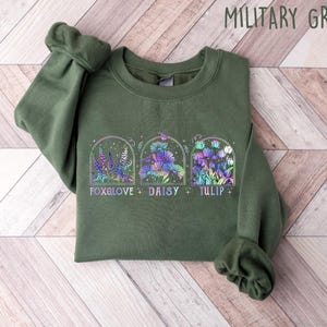Op de afbeelding: Een groen sweatshirt met een afbeelding van drie bloemen in bogen. De bloemen zijn een vingerhoedskruid, madeliefje en tulp. De tekst "Foxglove + Daisy + Tulip" staat onder de bloemen gedrukt.