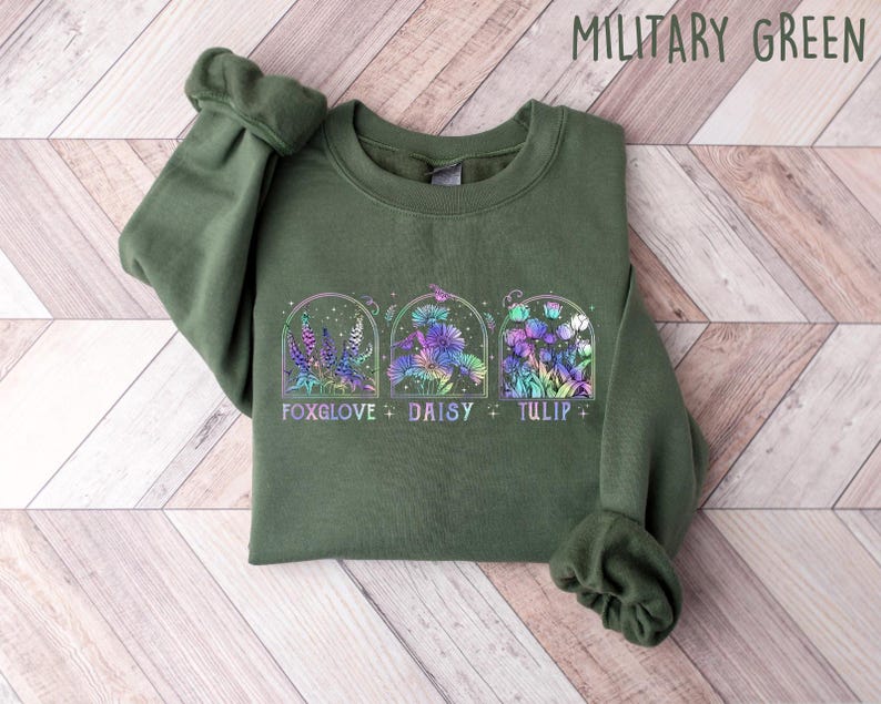 Op de afbeelding: Een groen sweatshirt met een afbeelding van drie bloemen in bogen. De bloemen zijn een vingerhoedskruid, madeliefje en tulp. De tekst "Foxglove + Daisy + Tulip" staat onder de bloemen gedrukt.