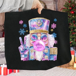 May include: Black graphic featuring a whimsical nutcracker with a peace sign and a bubblegum bubble. The nutcracker wears a blue and purple hat and jacket, with a gold band and accents. Blue and purple snowflakes surround the image.