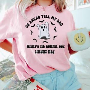Puede incluir: Una sudadera rosa claro con el texto "GO AHEAD TELL MY DAD WHAT'S HE GONNA DO? HAUNT ME?" y un gr&aacute;fico de fantasma. La sudadera se combina con pantalones cortos azul claro y una taza blanca con una carita sonriente amarilla.