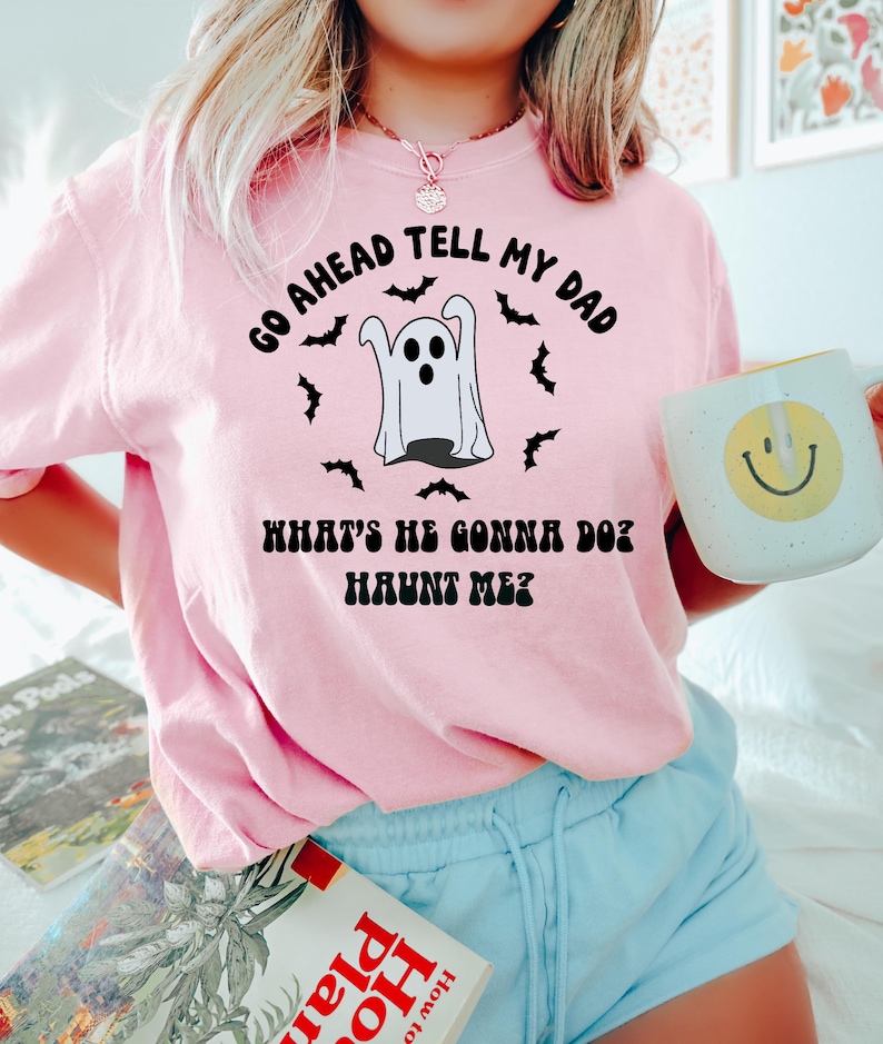 Puede incluir: Una sudadera rosa claro con el texto "GO AHEAD TELL MY DAD WHAT'S HE GONNA DO? HAUNT ME?" y un gr&aacute;fico de fantasma. La sudadera se combina con pantalones cortos azul claro y una taza blanca con una carita sonriente amarilla.
