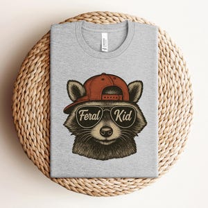May include: A heather gray t-shirt featuring a raccoon wearing a red baseball cap and sunglasses. The sunglasses have the words "Feral Kid" printed on them. The shirt is displayed on a woven, circular surface.