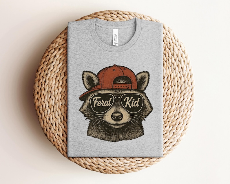 May include: A heather gray t-shirt featuring a raccoon wearing a red baseball cap and sunglasses. The sunglasses have the words "Feral Kid" printed on them. The shirt is displayed on a woven, circular surface.