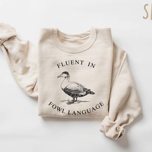 May include: A beige sweatshirt with the text "Fluent in Fowl Language" and a black and white illustration of a duck.