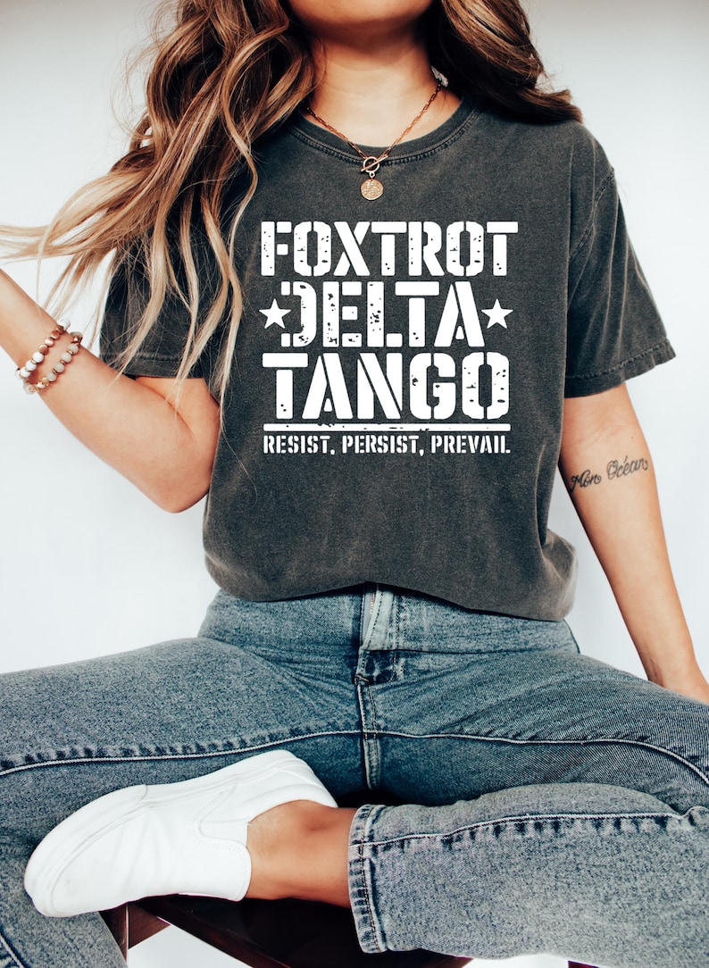 May include: Dark gray t-shirt with the words "FOXTROT DELTA TANGO" in large white block letters, with stars and the words "RESIST. PERSIST. PREVAIL." below. The shirt is paired with blue jeans and white slip-on shoes.