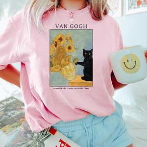 May include: Pink sweatshirt featuring a framed print of Van Gogh's "Sunflowers (Third Version) - 1888" with a black cat. The sweatshirt has the text "VAN GOGH" above the artwork. A white mug with a yellow smiley face is held in the hand.