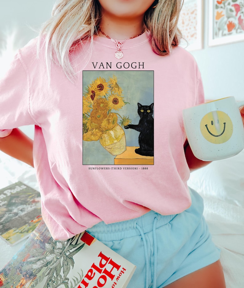 May include: Pink sweatshirt featuring a framed print of Van Gogh's "Sunflowers (Third Version) - 1888" with a black cat. The sweatshirt has the text "VAN GOGH" above the artwork. A white mug with a yellow smiley face is held in the hand.