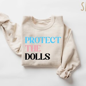 May include: A beige sweatshirt with the text "PROTECT THE DOLLS" printed on the front. The words "PROTECT" and "DOLLS" are in black, and the word "THE" is in pink.