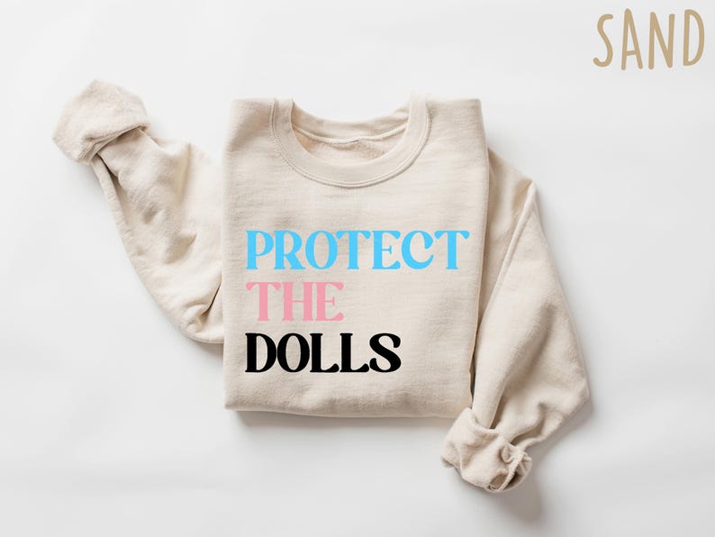 May include: A beige sweatshirt with the text "PROTECT THE DOLLS" printed on the front. The words "PROTECT" and "DOLLS" are in black, and the word "THE" is in pink.