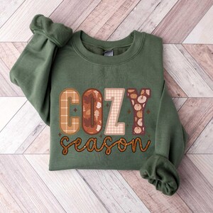 May include: A green sweatshirt with the text "cozy season" in brown, orange, and white patterned letters. The letters are decorated with fall-themed designs like leaves and pumpkins.