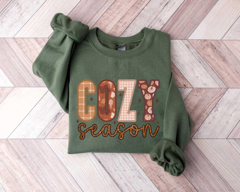May include: A green sweatshirt with the text "cozy season" in brown, orange, and white patterned letters. The letters are decorated with fall-themed designs like leaves and pumpkins.