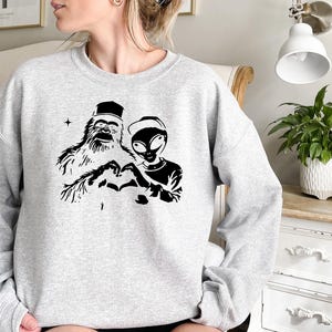 Puede incluir: Una sudadera gris con un estampado gr&aacute;fico negro de un yeti y un extraterrestre haciendo un coraz&oacute;n con sus manos.