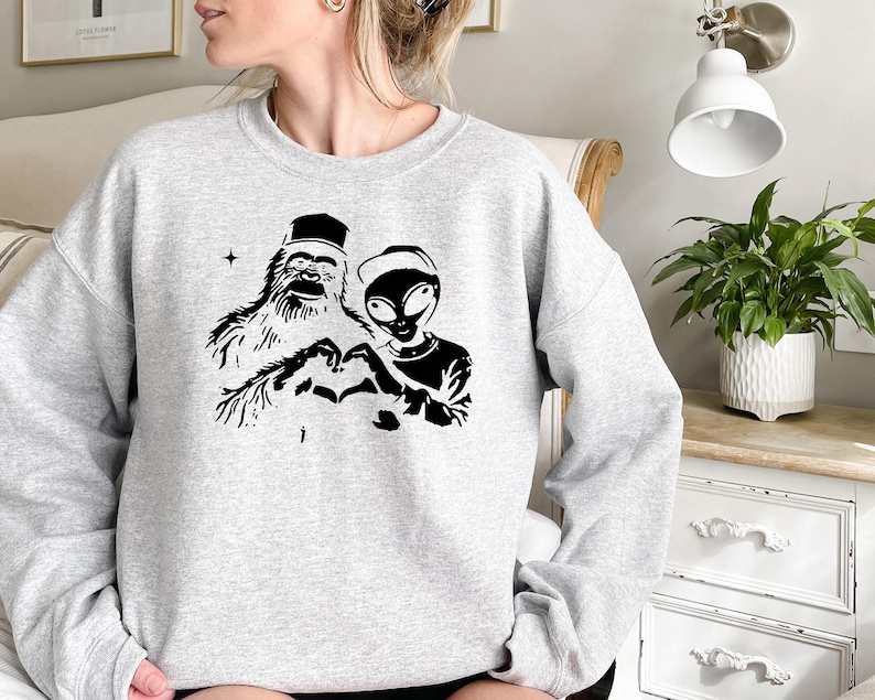 Puede incluir: Una sudadera gris con un estampado gr&aacute;fico negro de un yeti y un extraterrestre haciendo un coraz&oacute;n con sus manos.
