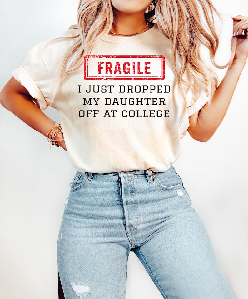 May include: Cream-colored t-shirt with the text "FRAGILE" in a red rectangle, and the phrase "I JUST DROPPED MY DAUGHTER OFF AT COLLEGE" in black. The person is wearing light blue jeans.