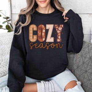 May include: A black sweatshirt with a graphic print that says "Cozy Season" in a patchwork style with fall colors and textures.