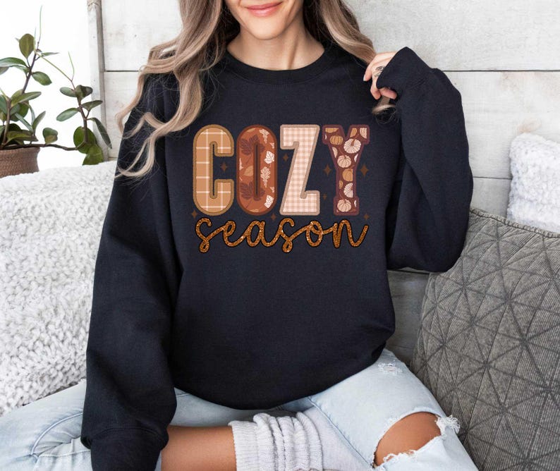 May include: A black sweatshirt with a graphic print that says "Cozy Season" in a patchwork style with fall colors and textures.