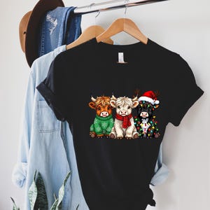 May include: Black t-shirt featuring a graphic of three Highland cows. One cow wears a green sweater, another a red scarf, and the third a Santa hat and Christmas lights. The shirt is hanging on a wooden hanger.