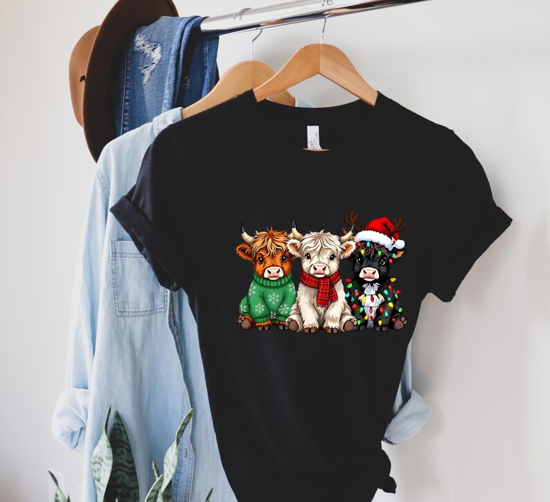 May include: Black t-shirt featuring a graphic of three Highland cows. One cow wears a green sweater, another a red scarf, and the third a Santa hat and Christmas lights. The shirt is hanging on a wooden hanger.