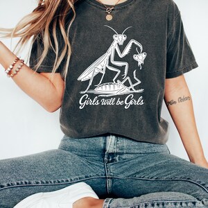 May include: Dark gray t-shirt featuring a white graphic of a praying mantis with a butterfly and the text "Girls will be Girls". The shirt is paired with blue jeans and white slip-on shoes.