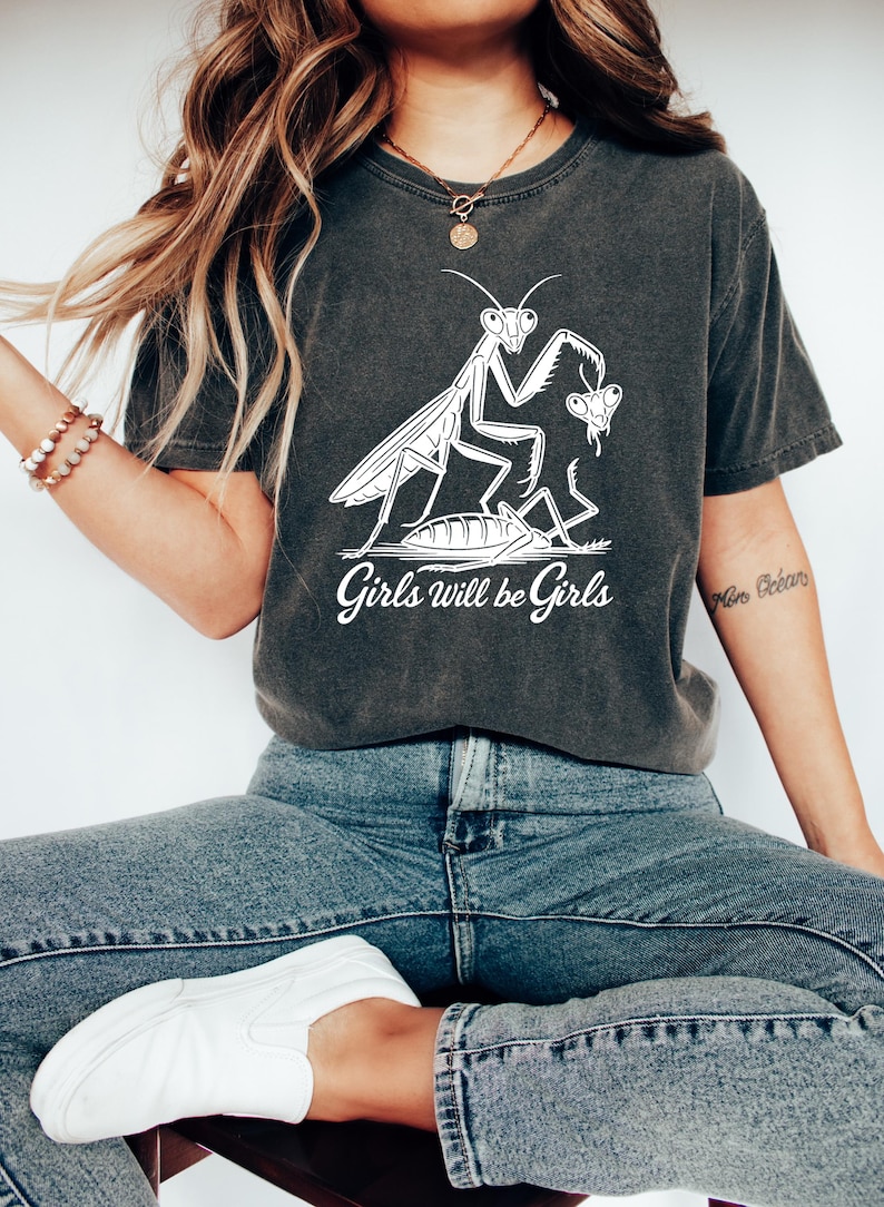 May include: Dark gray t-shirt featuring a white graphic of a praying mantis with a butterfly and the text "Girls will be Girls". The shirt is paired with blue jeans and white slip-on shoes.