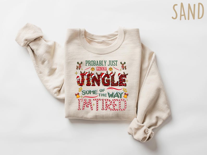 May include: A sand-colored sweatshirt with the text "Probably Just Gonna Jingle Some of the Way I'm Tired" in a festive design. The word "Jingle" is in a plaid pattern. The sweatshirt has long sleeves.