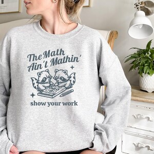 May include: Gray sweatshirt with a graphic of two raccoons using a calculator. The text reads "The Math Ain't Mathin' show your work."