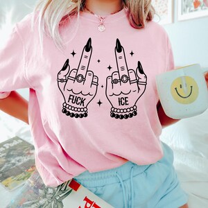 May include: Pink crew neck sweatshirt with a black graphic of two hands displaying the middle finger, with the words "FUCK ICE". The hands are adorned with rings and bracelets. A white mug with a yellow smiley face is held in the right hand.