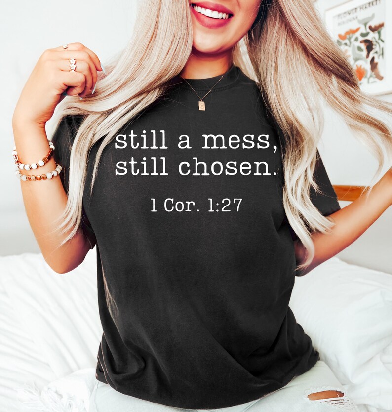 K&ouml;nnte beinhalten: Schwarzes T-Shirt mit dem wei&szlig;en Text "still a mess, still chosen." und dem Bibelvers 1 Kor. 1:27. Das Shirt hat einen Rundhalsausschnitt.