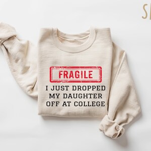 May include: A sand-colored sweatshirt with the text "FRAGILE" in a red rectangle, and the phrase "I JUST DROPPED MY DAUGHTER OFF AT COLLEGE" in black. The sleeves are folded.