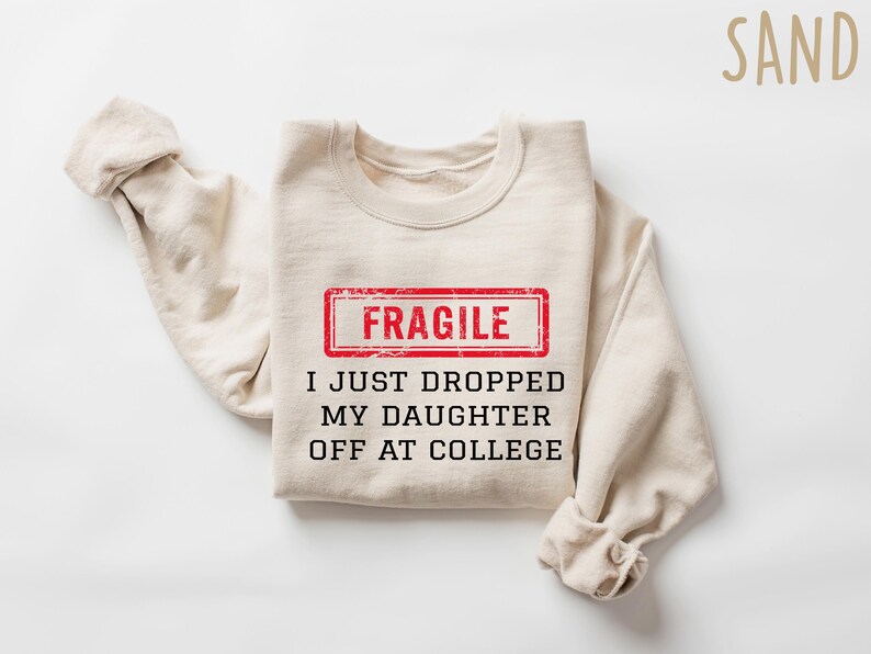 May include: A sand-colored sweatshirt with the text "FRAGILE" in a red rectangle, and the phrase "I JUST DROPPED MY DAUGHTER OFF AT COLLEGE" in black. The sleeves are folded.