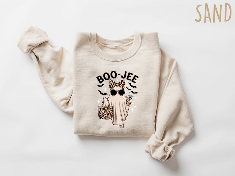 May include: Sand-colored sweatshirt with a graphic of a ghost wearing sunglasses and a leopard print bow, holding a leopard print tote bag and a drink. The text "BOO-JEE" and bats are above the ghost. The word "SAND" is in the upper right corner.