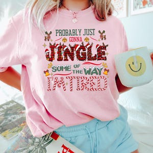 May include: A light pink sweatshirt with the text "Probably Just Gonna Jingle Some of the Way I'm Tired" in a festive Christmas design. The word "Jingle" is in a plaid pattern. The person is holding a white mug with a yellow smiley face.