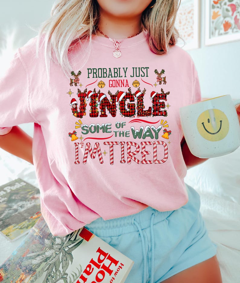 May include: A light pink sweatshirt with the text "Probably Just Gonna Jingle Some of the Way I'm Tired" in a festive Christmas design. The word "Jingle" is in a plaid pattern. The person is holding a white mug with a yellow smiley face.