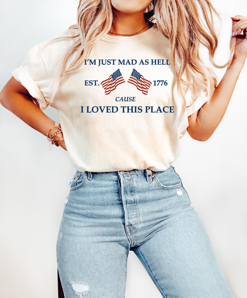 May include: Cream-colored t-shirt with the text "I'M JUST MAD AS HELL CAUSE I LOVED THIS PLACE" and two American flags. The shirt is paired with light blue jeans.