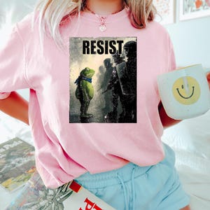 May include: Pink sweatshirt with a graphic print featuring a frog wearing a blue scarf facing a line of silhouetted figures. The word "RESIST" is printed above the frog. A white mug with a smiley face is held in the hand.