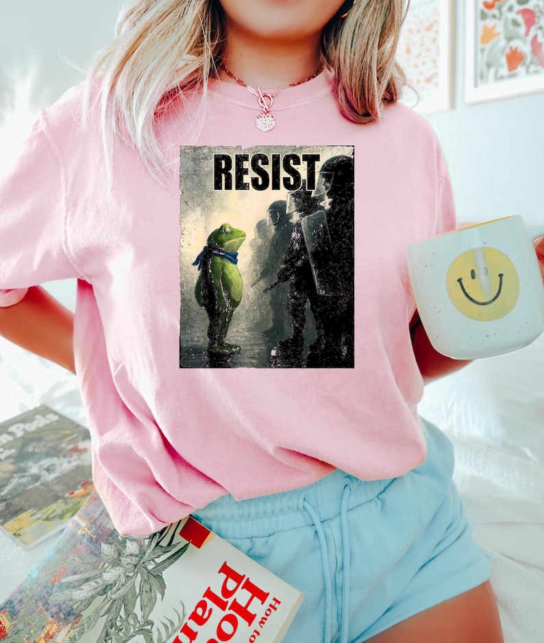 May include: Pink sweatshirt with a graphic print featuring a frog wearing a blue scarf facing a line of silhouetted figures. The word "RESIST" is printed above the frog. A white mug with a smiley face is held in the hand.