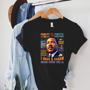 K&ouml;nnte beinhalten: Schwarzes T-Shirt mit einem Portr&auml;t von Martin Luther King Jr. und dem Text "I Have a Dream". Das Design enth&auml;lt S&auml;tze wie "Gleichheit f&uuml;r alle", "Menschenrechte" und "B&uuml;rgerrechte". Das T-Shirt h&auml;ngt an einem Holzb&uuml;gel.