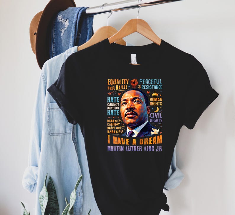 K&ouml;nnte beinhalten: Schwarzes T-Shirt mit einem Portr&auml;t von Martin Luther King Jr. und dem Text "I Have a Dream". Das Design enth&auml;lt S&auml;tze wie "Gleichheit f&uuml;r alle", "Menschenrechte" und "B&uuml;rgerrechte". Das T-Shirt h&auml;ngt an einem Holzb&uuml;gel.