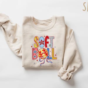 Puede incluir: Una sudadera beige con las palabras "SOFT BALL" en letras coloridas y estampadas. Las letras est&aacute;n decoradas con estrellas, rayas y un gr&aacute;fico de softbol.