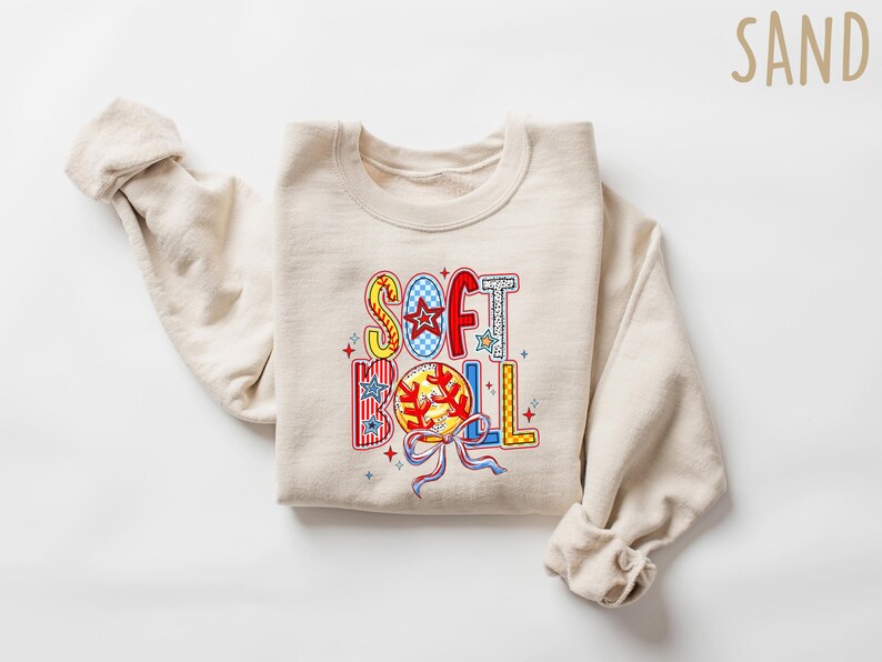 Puede incluir: Una sudadera beige con las palabras "SOFT BALL" en letras coloridas y estampadas. Las letras est&aacute;n decoradas con estrellas, rayas y un gr&aacute;fico de softbol.