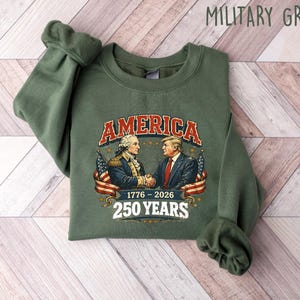 Puede incluir: Sudadera verde militar con un gr&aacute;fico de George Washington y Donald Trump d&aacute;ndose la mano, la palabra "AMERICA" en una fuente vintage y el texto "1776-2026 250 YEARS". La sudadera est&aacute; sobre una superficie de madera clara.