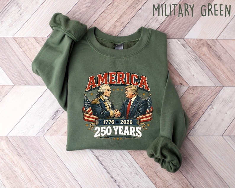 Puede incluir: Sudadera verde militar con un gr&aacute;fico de George Washington y Donald Trump d&aacute;ndose la mano, la palabra "AMERICA" en una fuente vintage y el texto "1776-2026 250 YEARS". La sudadera est&aacute; sobre una superficie de madera clara.