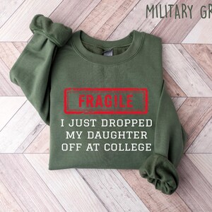 May include: Military green sweatshirt with the word "FRAGILE" in a red rectangle, and the text "I JUST DROPPED MY DAUGHTER OFF AT COLLEGE" in white. The sweatshirt is folded on a light wood-grain surface.