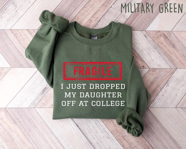 May include: Military green sweatshirt with the word "FRAGILE" in a red rectangle, and the text "I JUST DROPPED MY DAUGHTER OFF AT COLLEGE" in white. The sweatshirt is folded on a light wood-grain surface.