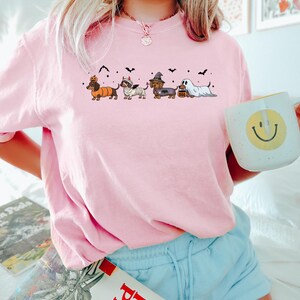 May include: A light pink t-shirt featuring a Halloween-themed dachshund design. The graphic shows five dachshunds dressed in costumes, including a pumpkin, mummy, witch, and ghost. Black bats are scattered above the dogs. The person is holding a white mug with a yellow smiley face.