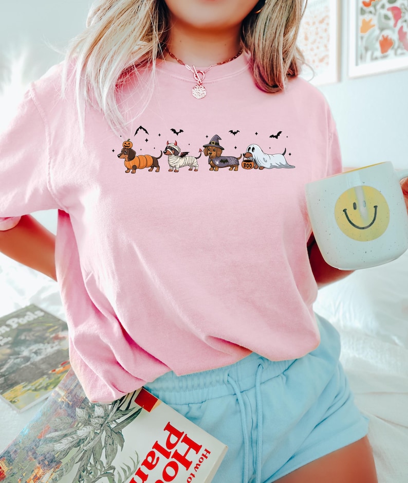 May include: A light pink t-shirt featuring a Halloween-themed dachshund design. The graphic shows five dachshunds dressed in costumes, including a pumpkin, mummy, witch, and ghost. Black bats are scattered above the dogs. The person is holding a white mug with a yellow smiley face.