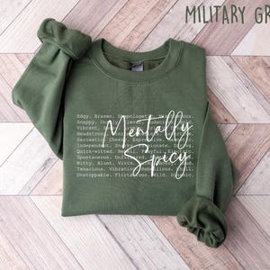 May include: A military green sweatshirt with the words "Mentally Spicy" in a white, cursive font. The sweatshirt has a crew neck and long sleeves. The text includes a list of adjectives such as "edgy", "brazen", and "unfiltered".