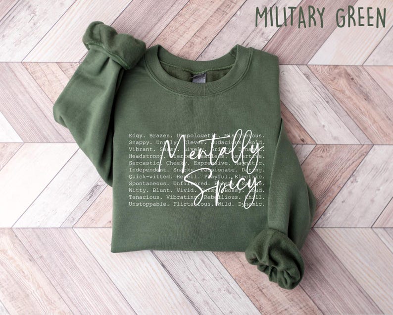May include: A military green sweatshirt with the words "Mentally Spicy" in a white, cursive font. The sweatshirt has a crew neck and long sleeves. The text includes a list of adjectives such as "edgy", "brazen", and "unfiltered".
