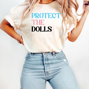 May include: A white t-shirt with the text "PROTECT THE DOLLS" in blue and pink letters.