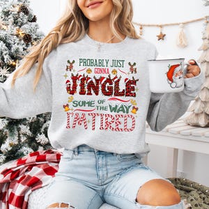May include: A light gray sweatshirt with the text "Probably Just Gonna Jingle Some of the Way I'm Tired" in red, green, and black. The sweatshirt features Christmas-themed graphics. A white mug with a Santa Claus design is held in the hand.